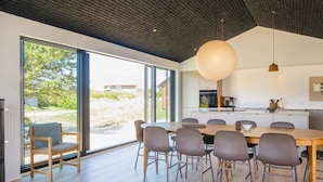 A modern kitchen with a large island, a dining area with a wooden table and chairs, and large sliding glass doors leading to a garden. - 5 Star Holiday Home in Romo (Rømø)