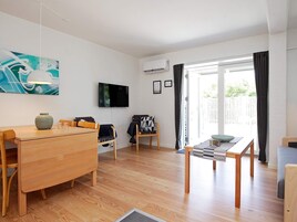 Ein modernes Wohnzimmer mit einem Holzesstisch, zwei Stühlen, einem an der Wand montierten Fernseher und einem großen Fenster mit Vorhängen.