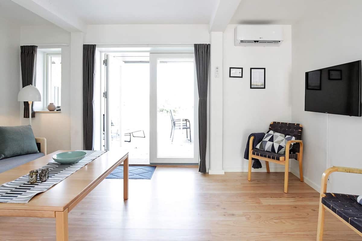 Un salon moderne avec un plancher en bois, un canapé, une table à manger avec un saladier et des verres, un téléviseur et un fauteuil avec un coussin.