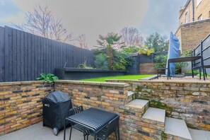 A backyard with a brick wall, a barbecue grill, a table, and a covered seating area. - Contemporary 2BD Cottage-Style Flat, Hackney (Greater London)