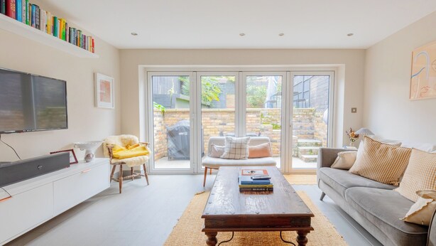 A living room with a flat-screen TV, a coffee table with books, a sofa, and a chair. There is a bookshelf with books and a large window with a view of the outside. - Contemporary 2BD Cottage-Style Flat, Hackney (Greater London)