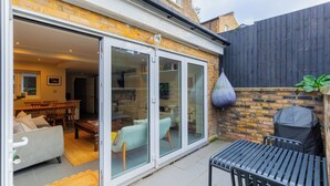 A patio area with a sliding glass door leading to an indoor living space, a barbecue grill, and a dining area with a table and chairs. - Contemporary 2BD Cottage-Style Flat, Hackney (Greater London)