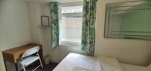 A small room with a bed, a desk, and a chair. There is a window with curtains, a radiator, and a framed notice on the wall.