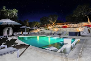 A nighttime swimming pool area with lounge chairs, umbrellas, and a stone wall.