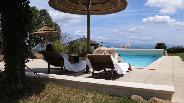 A poolside area with two lounge chairs and a table, a thatched umbrella, and a view of the pool and mountains.
