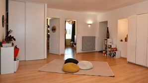 A spacious living room with a wooden floor, a large rug with three cushions, a ceiling light fixture, and a bookshelf with books and decorative items. - Welcome to the" Domaine des étoiles"<br>Enjoy a moment of escape and fun! (GILLONNAY)