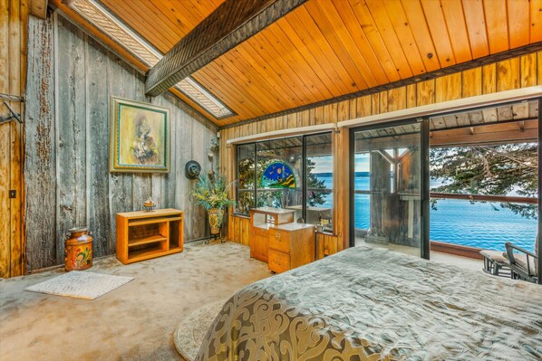 A bedroom with a large bed, wooden walls, a wooden ceiling, a window with a view of the sea, and a small wooden dresser. - Rita s Lodge in Brinnon (Brinnon)