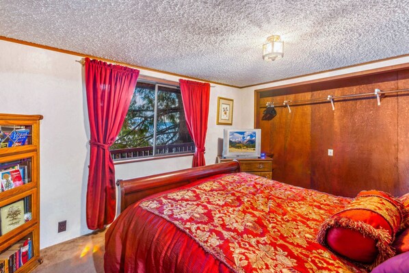 Ein Schlafzimmer mit einem großen Bett, einem Holzschrank, einem Bücherregal, einem Fenster mit roten Vorhängen und einer Deckenleuchte.
