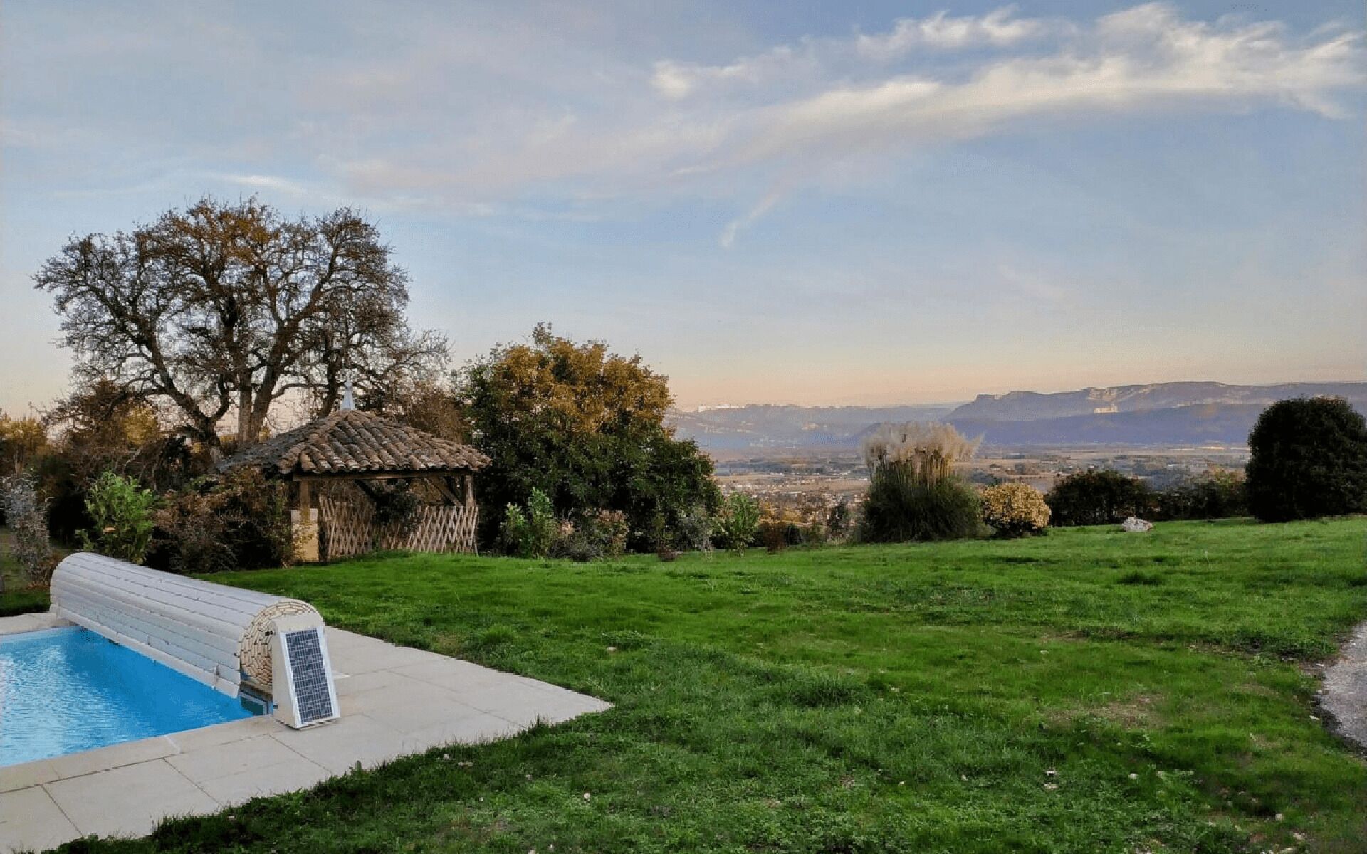 A swimming pool with a tiled deck, a small gazebo, and a grassy lawn with a view of distant mountains.