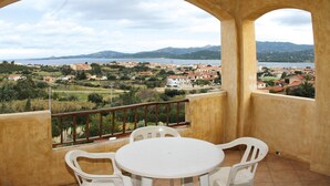 Un balcon avec une table et des chaises, donnant sur une ville côtière, avec des bâtiments et des montagnes visibles au loin.