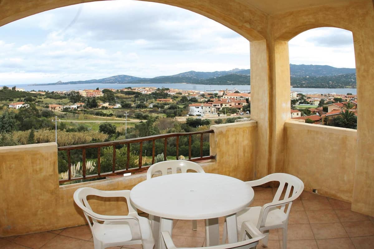 A balcony with a table and chairs, overlooking a coastal town with buildings and mountains in the distance.