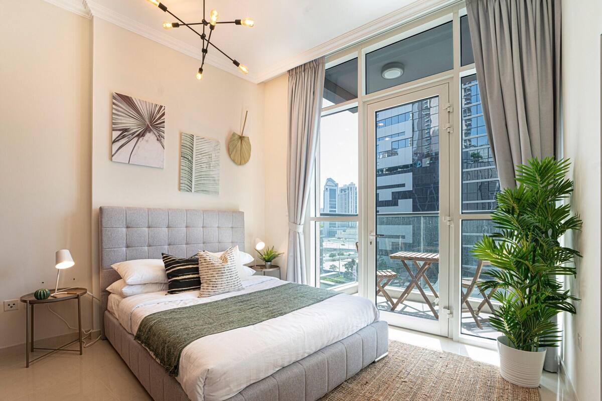 Quarto moderno com uma cama grande, vista da cidade através de janelas do chão ao teto e uma varanda com planta em vaso.