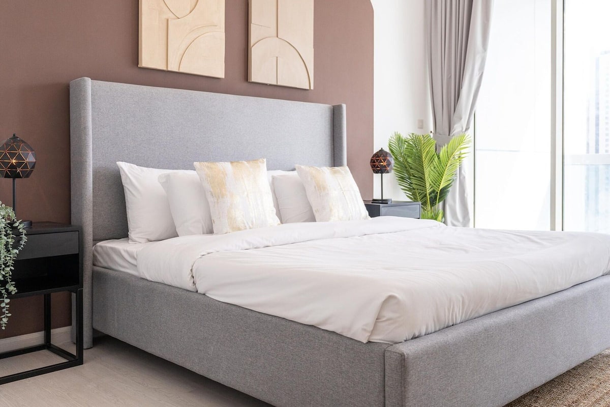 A modern bedroom with a grey upholstered bed, white bedding, and a grey headboard. There are two wall-mounted artworks above the bed and a potted plant to the right.