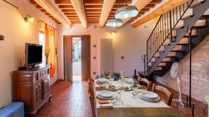 A dining area with a wooden table set for four, a television on a cabinet, and a staircase with a fan. - Apartment in a Farmhouse With 2 Swimming Pools (Cagli)