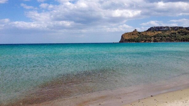 A beach with clear turquoise water, a sandy shore, and a rocky hill with a structure on top. - Apartment in a Farmhouse With 2 Swimming Pools (Cagli)