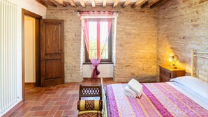 A bedroom with a stone wall, wooden beams, a bed with a pink and purple quilt, a wooden chair, and a window with a curtain. - Apartment in a Farmhouse With 2 Swimming Pools (Cagli)
