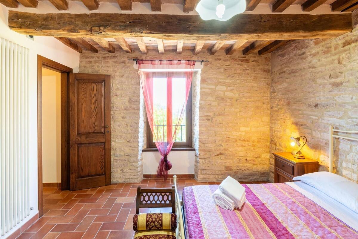 Une chambre avec un mur en pierre, des poutres en bois, un lit recouvert d’une couette rose et violette, une chaise en bois et une fenêtre avec un rideau.