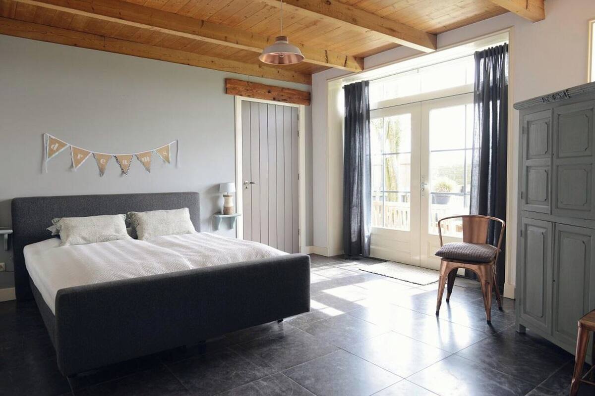 A bedroom with a large bed, a wooden ceiling, a window with curtains, and a chair.