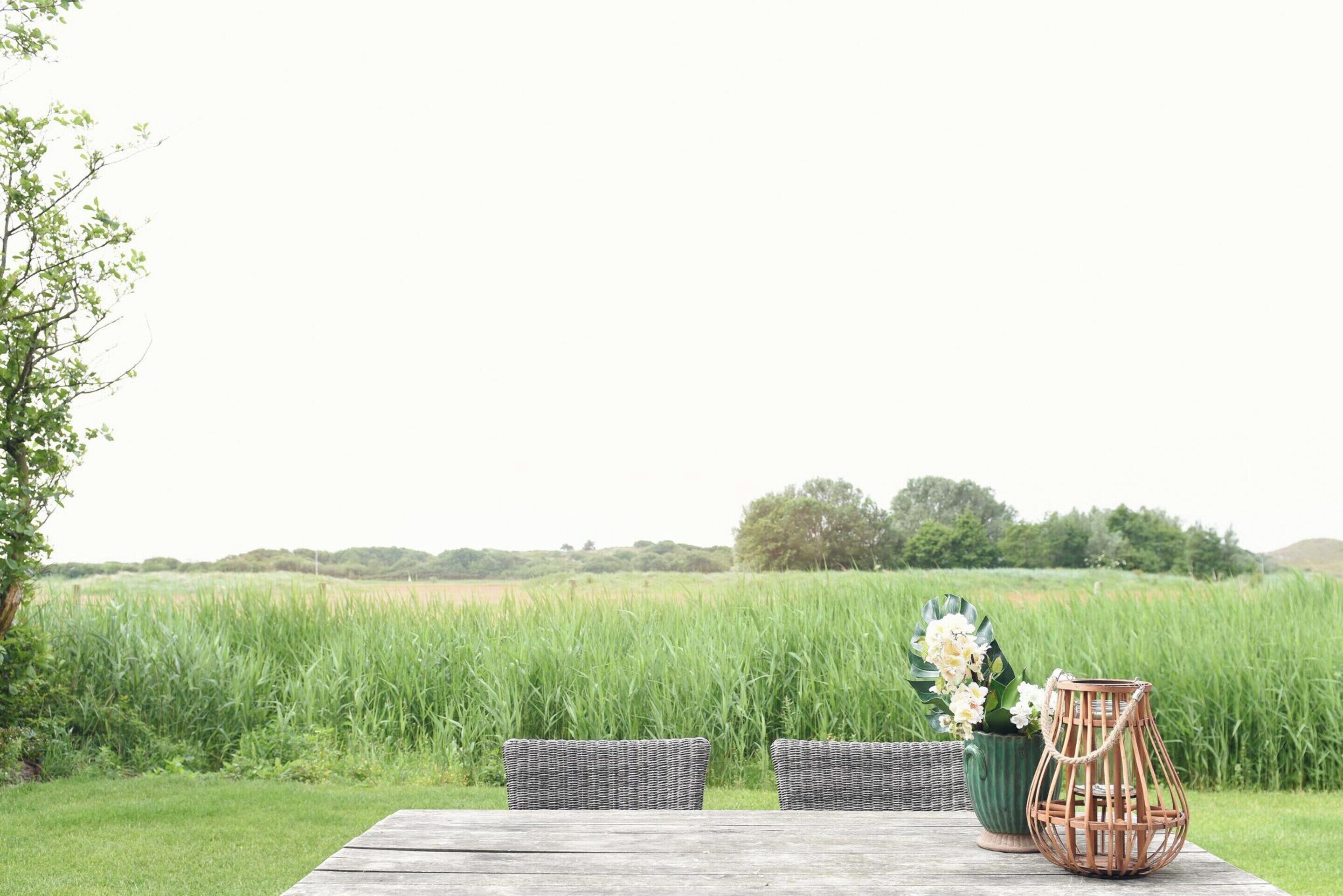 A wooden table with two wicker chairs and a wicker lantern in a grassy field with a view of a distant tree line.