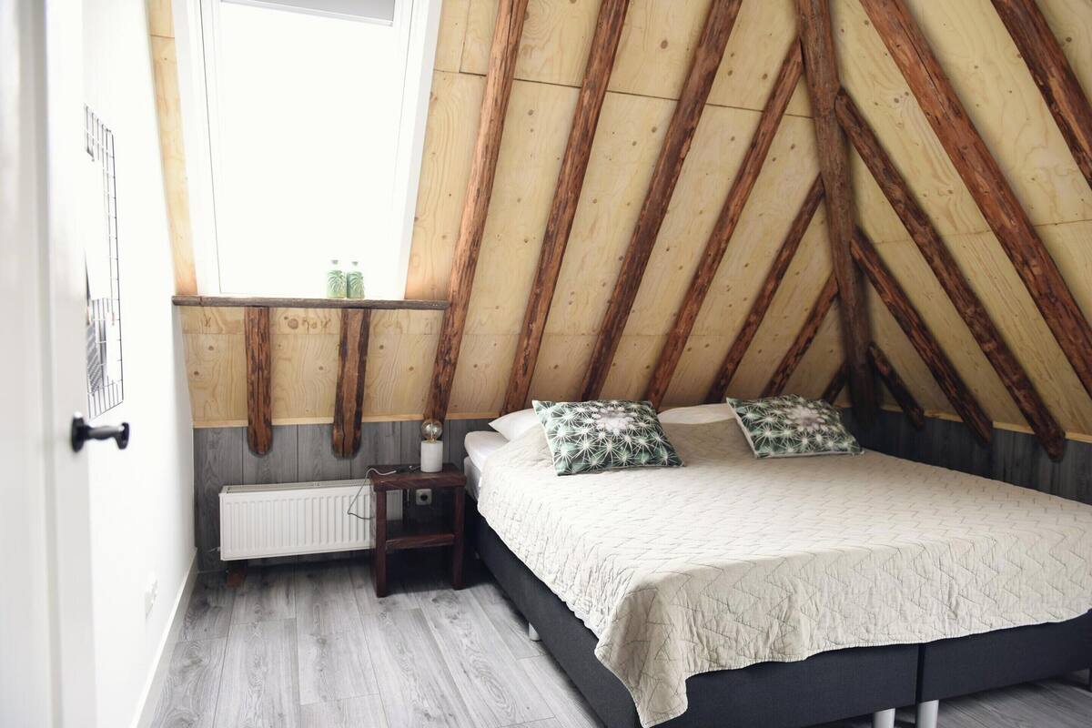 A bedroom with a sloped ceiling, a bed with a quilted cover, two pillows, a bedside table with a lamp, and a window with a view of the outside.