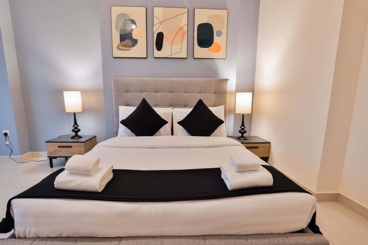 A neatly made bed with a grey upholstered headboard, two bedside tables with lamps, and abstract artwork on the wall.