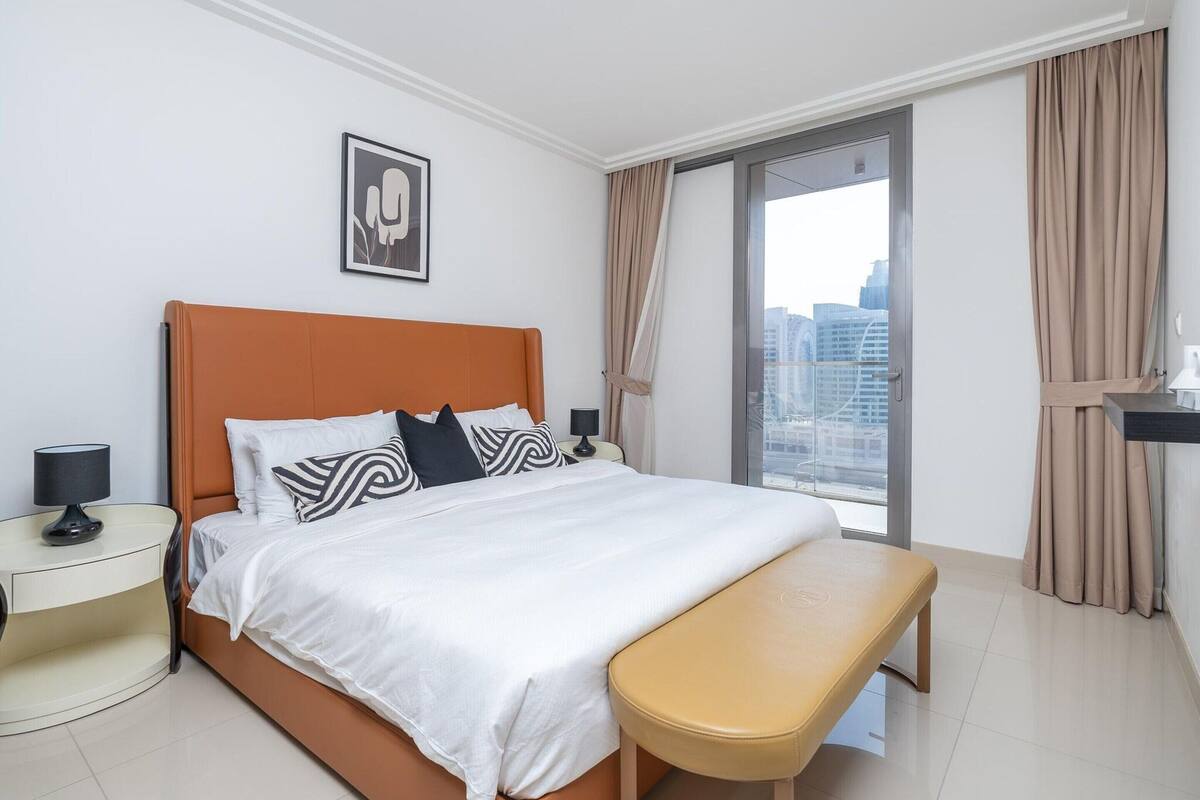 Ein modernes Hotelzimmer mit einem großen Bett, einer Bank, einem Nachttisch und Blick auf die Stadt durch ein großes Fenster.
