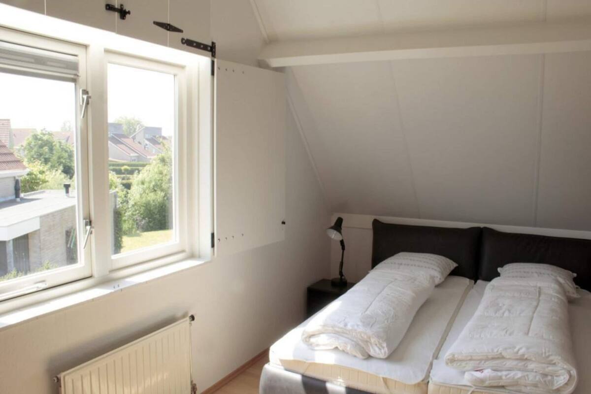 Ein Schlafzimmer mit Bett, Nachttisch, Lampe und einem Fenster mit Blick auf Häuser und Bäume.
