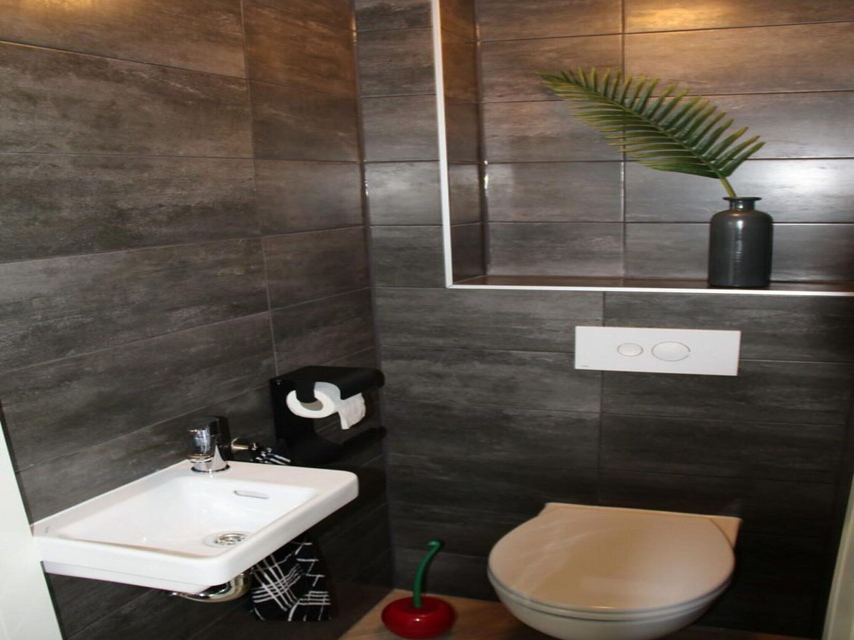A modern bathroom with dark tiles, a white sink, a toilet, and a mirror with a potted plant.