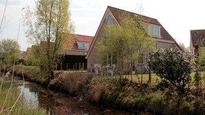 En bostadsbyggnad med ett sluttande tak, omgiven av grönska och en liten vattenyta.