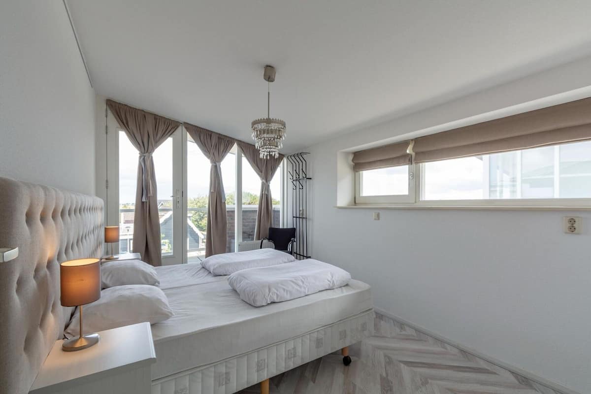 A bedroom with a bed, a bedside lamp, a chandelier, a window with curtains, and a view of the outside.