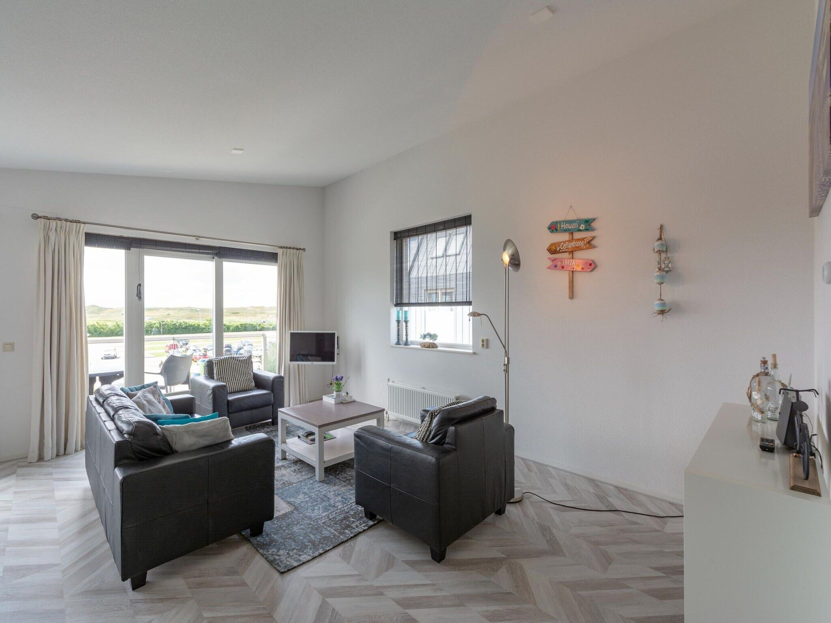 Ein modernes Wohnzimmer mit einer Sofagarnitur, einem Couchtisch und einem Fernseher. Ein Fenster mit Vorhängen, eine Stehlampe und eine Wanddekoration sind ebenfalls vorhanden.