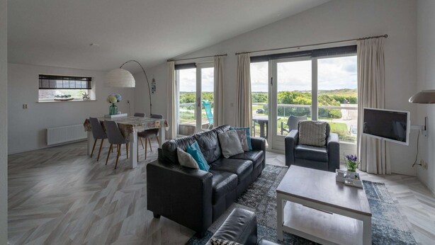 A spacious living area with a black leather sofa, a coffee table, and a TV. There is a dining area with a table and chairs, and large windows offering a view of the outdoors. - Fantastic Apartment Seaside With Terrace (Callantsoog)