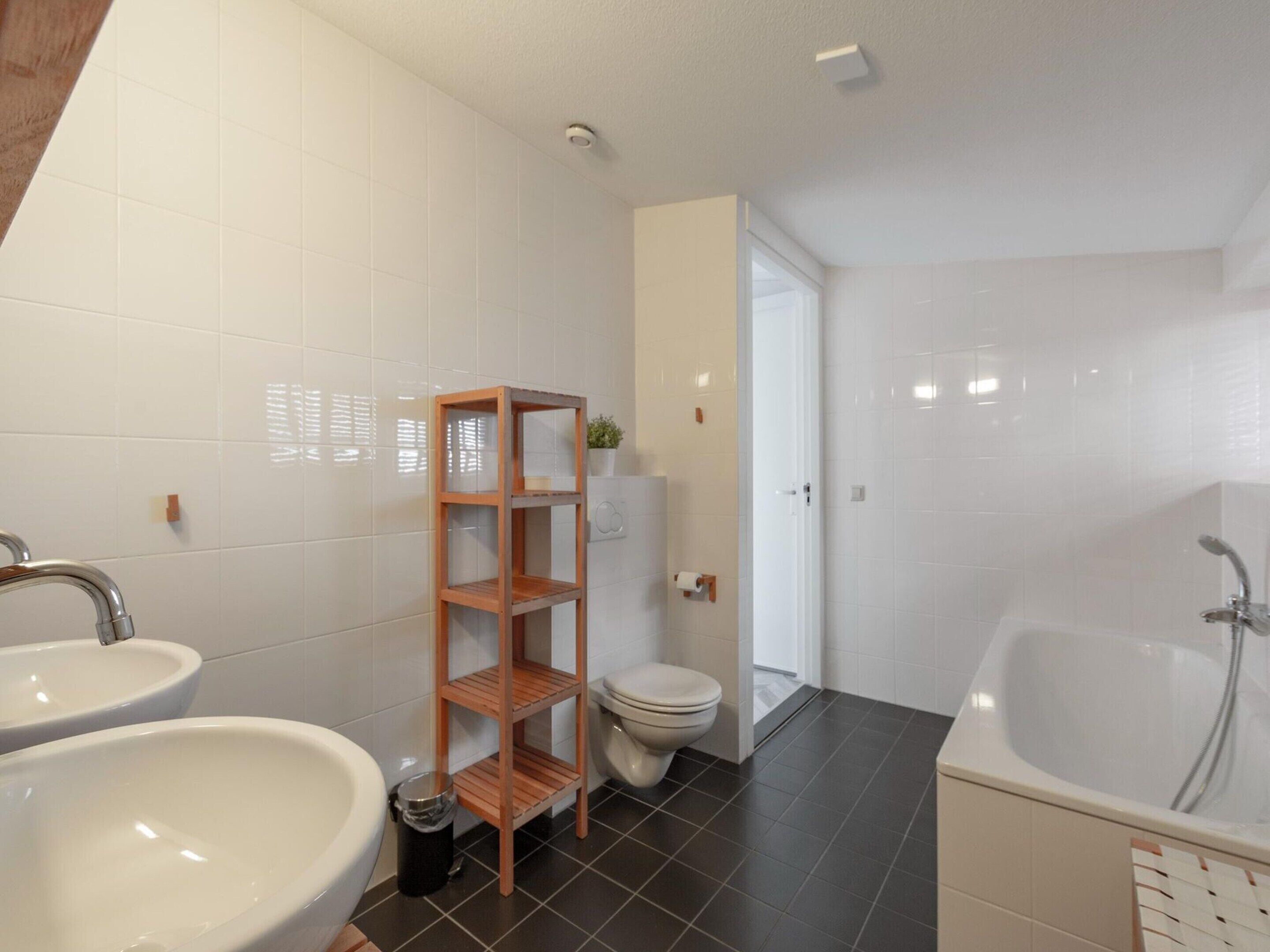 A bathroom with white tiles, a wooden shelf, a toilet, and a bathtub.