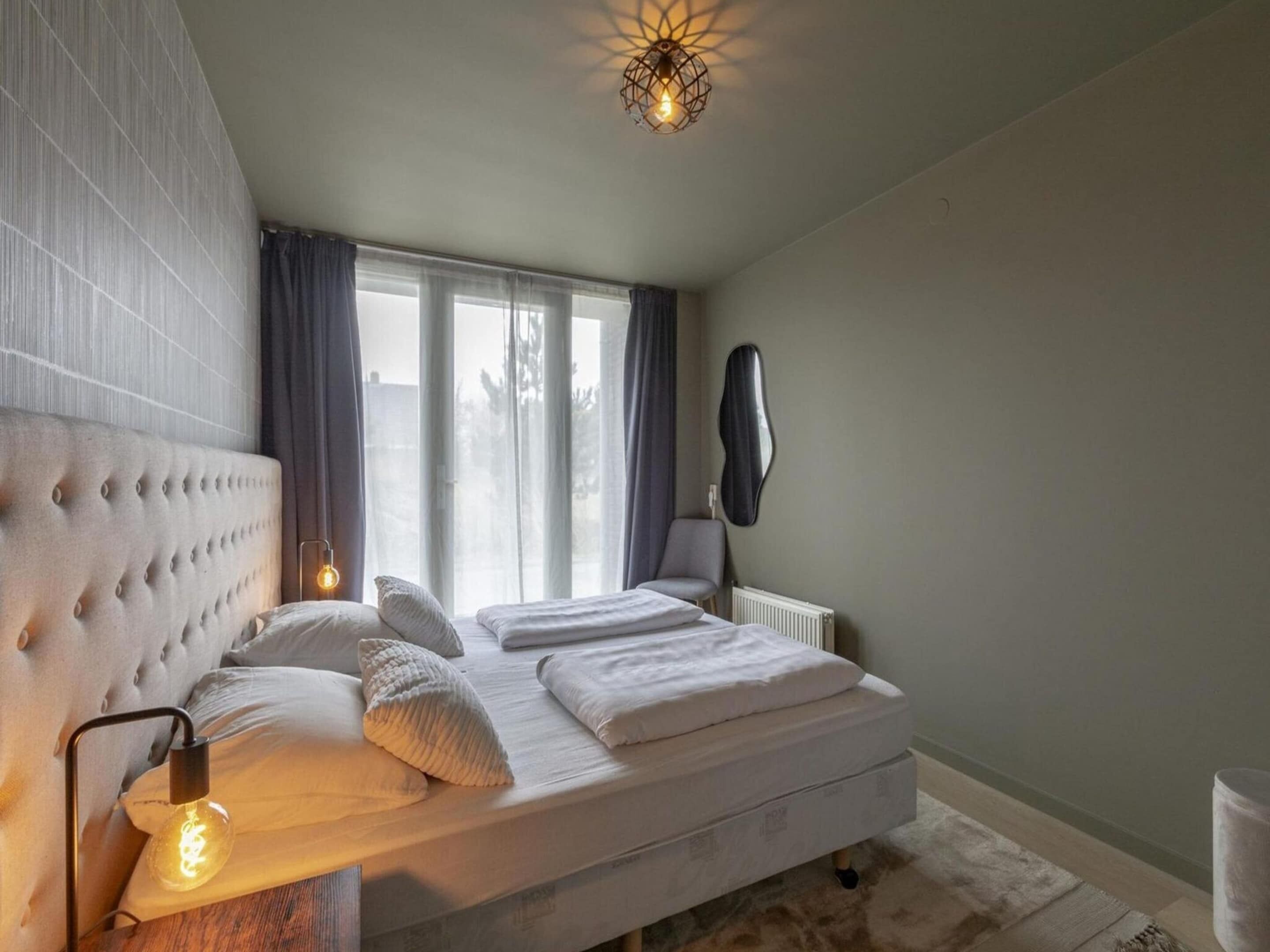 A bedroom with a tufted headboard, a bed with white bedding, a radiator, and a wall-mounted light fixture.