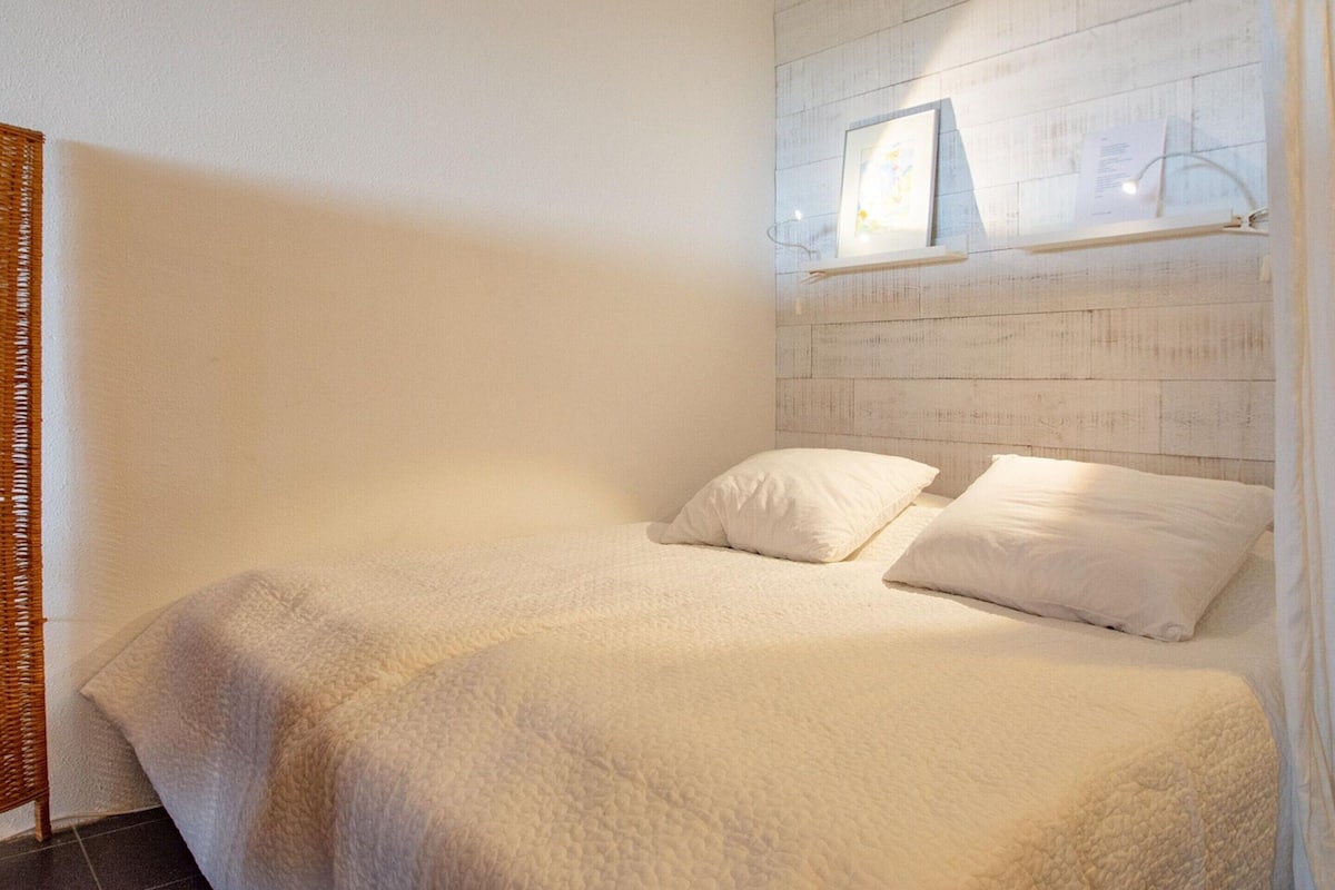 Un dormitorio con cama, almohadas, cabecera con estante y lámpara de pared.