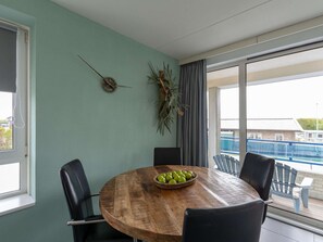 Een eetruimte met een houten tafel, vier stoelen, een fruitschaal en een uitzicht op een zwembad door grote ramen.