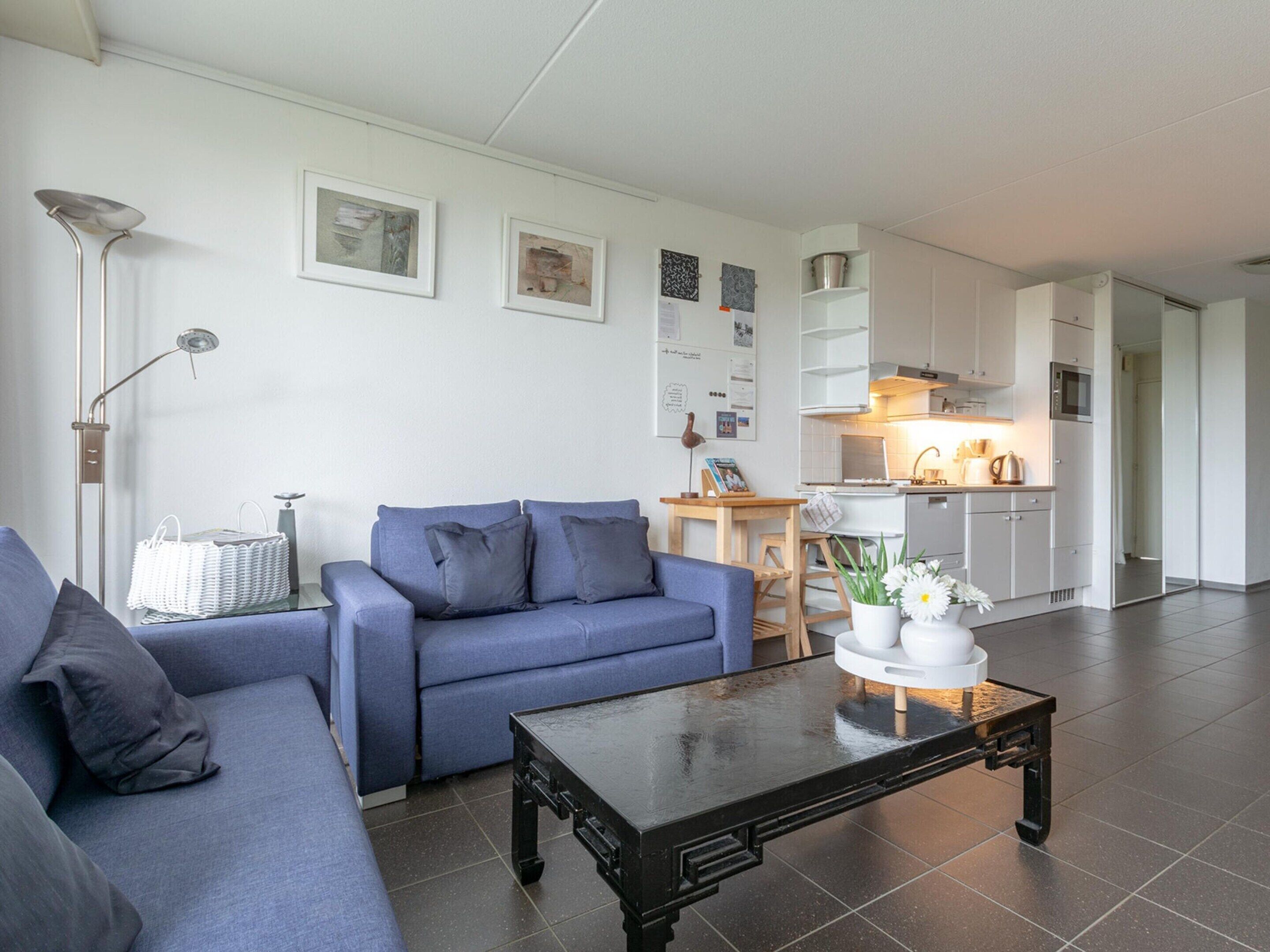 A modern living room with a blue sofa, a coffee table, and a kitchen area with white cabinets and a microwave.