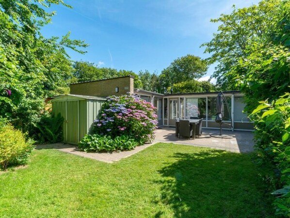 Un jardin à l’arrière d’une maison, avec un abri de jardin vert, une terrasse dotée d’une table et de chaises, et un grand arbuste aux fleurs violettes.