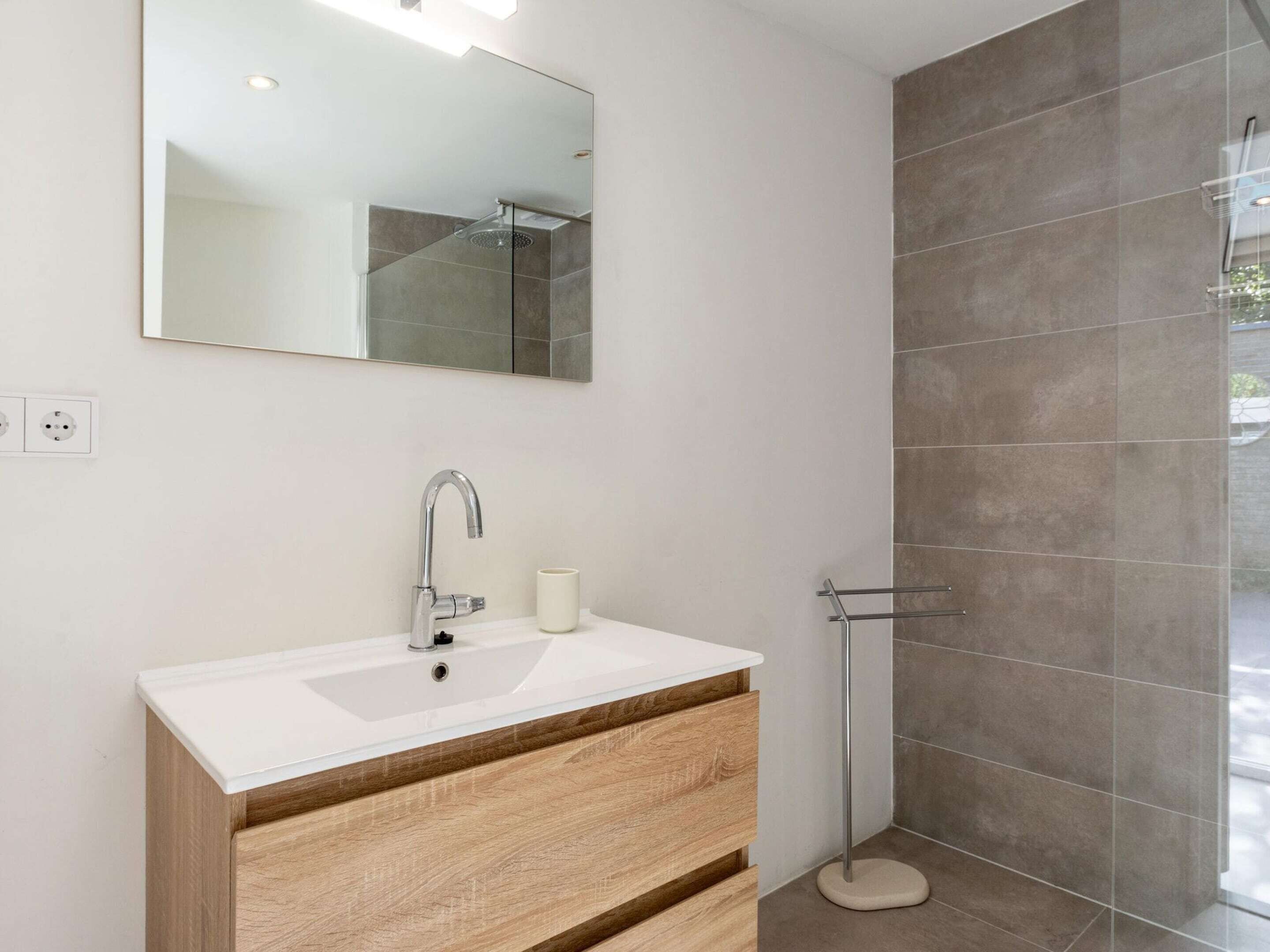 A modern bathroom with a white sink, wooden drawers, a mirror, and a glass shower enclosure.