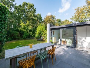 En moderne udendørs terrasse med et træbord og stole, omgivet af frodigt grønt og en moderne bygning med glasfacade.