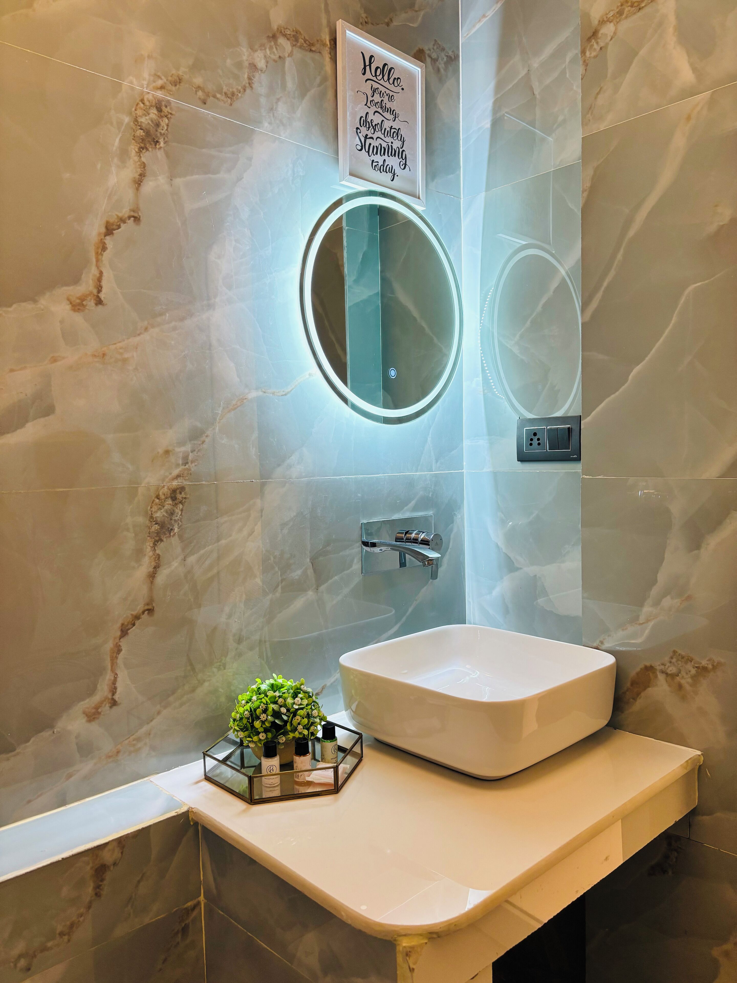 Un baño con pared de mármol, un espejo redondo, un lavabo blanco y una planta pequeña en la encimera.