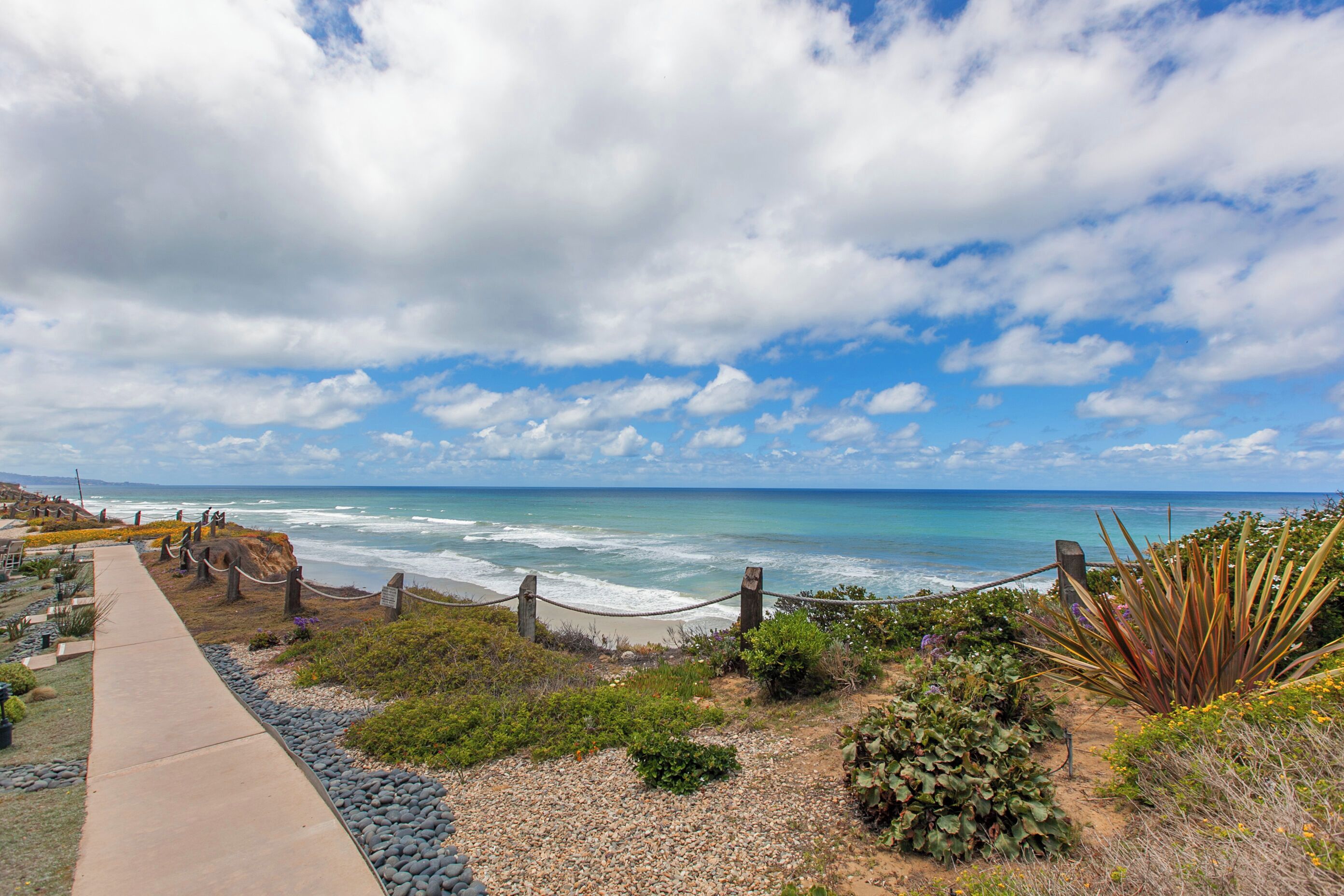 A coastal walkway with a view of the ocean, a sandy beach, and a clear sky.