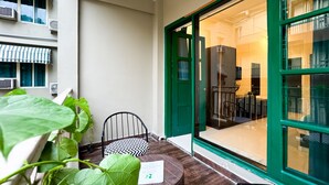Un balcón pequeño con una mesa y sillas, una planta verde grande y vistas al interior del edificio.