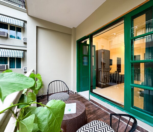 Un balcón pequeño con una mesa y sillas, una planta verde grande y vistas al interior del edificio.