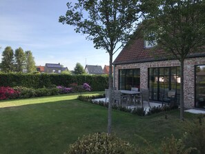 Ein Wohnhaus mit Backsteinfassade, einer überdachten Terrasse mit Gartenmöbeln und einem gepflegten Garten mit Hecken und blühenden Pflanzen.