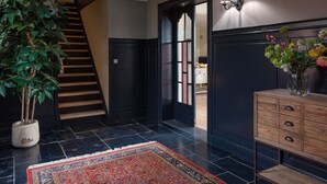 Ein modernes Interieur mit einer dunklen Holztreppe, einer großen Topfpflanze, einem gemusterten Teppich und einem Holzschrank mit einer Blumenvase.