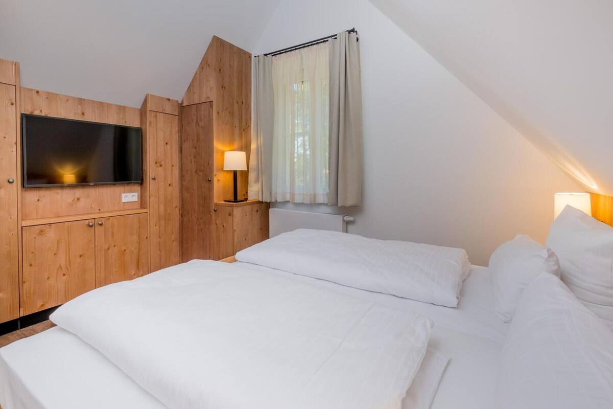 A bedroom with a sloped ceiling, wooden paneling, a TV mounted on the wall, two bedside lamps, and a bed with white bedding.