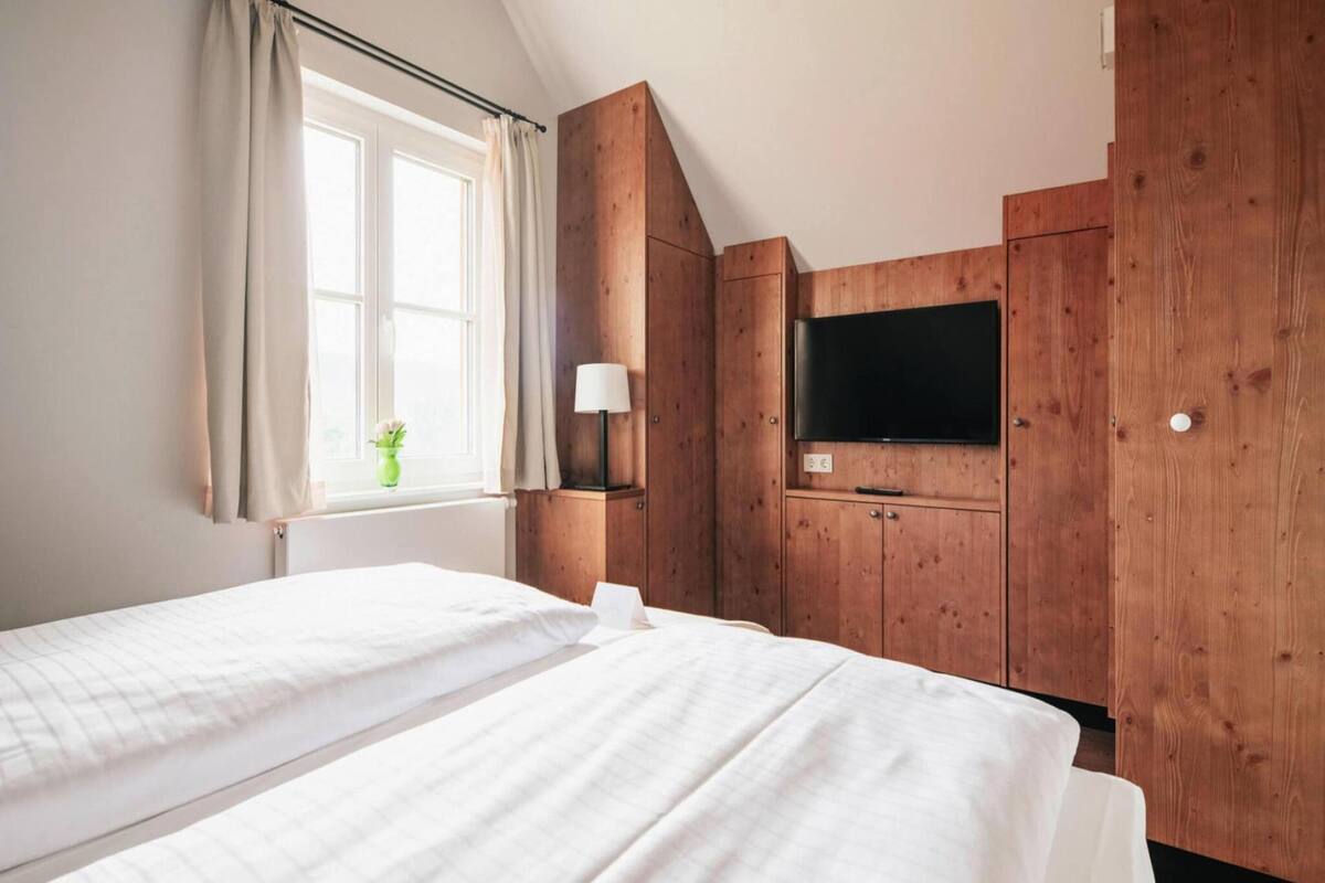 A room with two beds, a TV, wooden cabinets, a window with curtains, and a small plant.