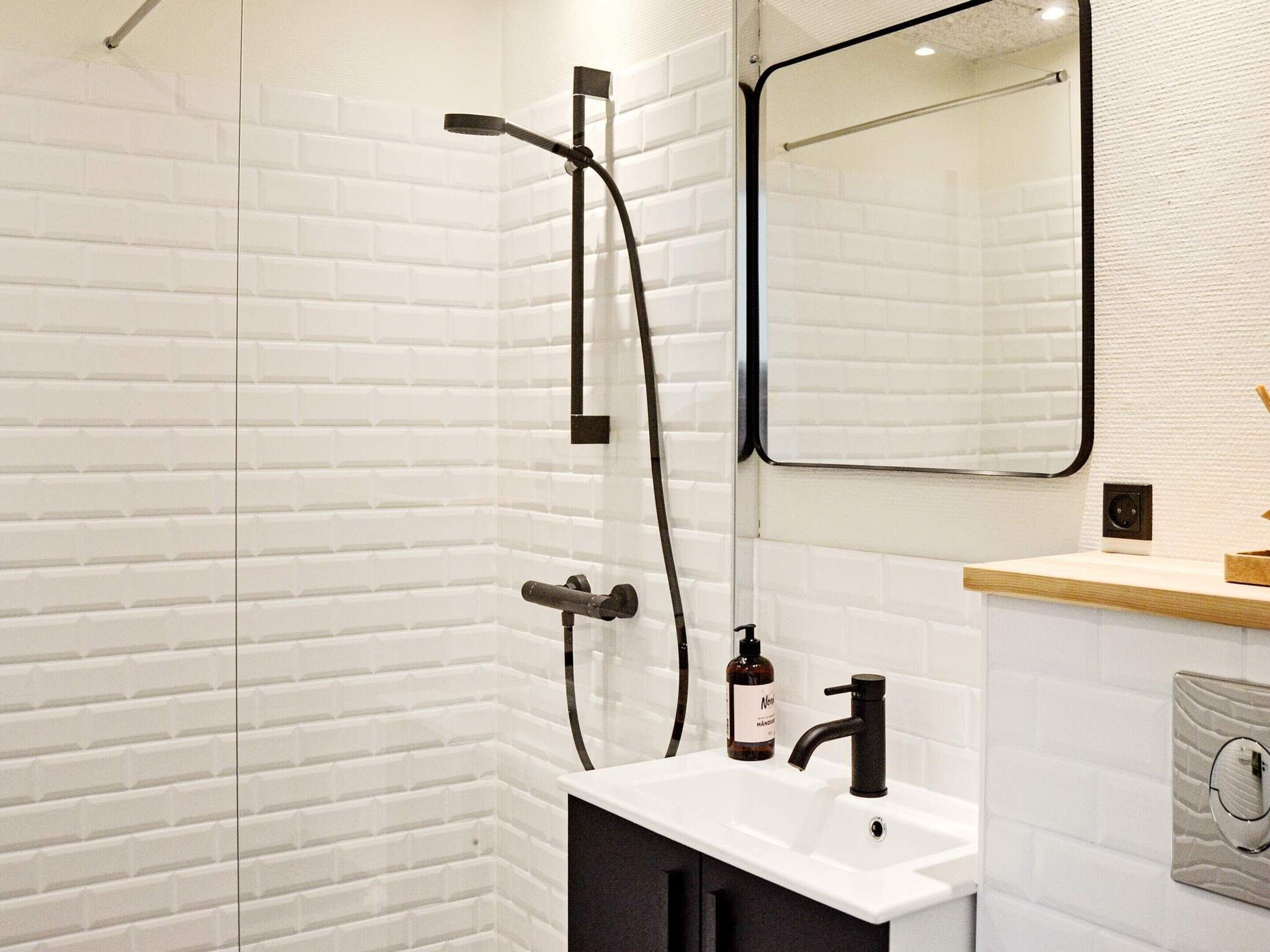 A modern bathroom with a white brick wall, a large mirror, a shower with a handheld showerhead, a sink with a black faucet, and a soap dispenser.