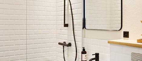 A modern bathroom with a white brick wall, a large mirror, a shower with a handheld showerhead, a sink with a black faucet, and a soap dispenser.